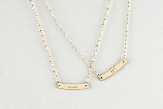 Two gold necklaces with personalized gold bar pendants on a light gray background
