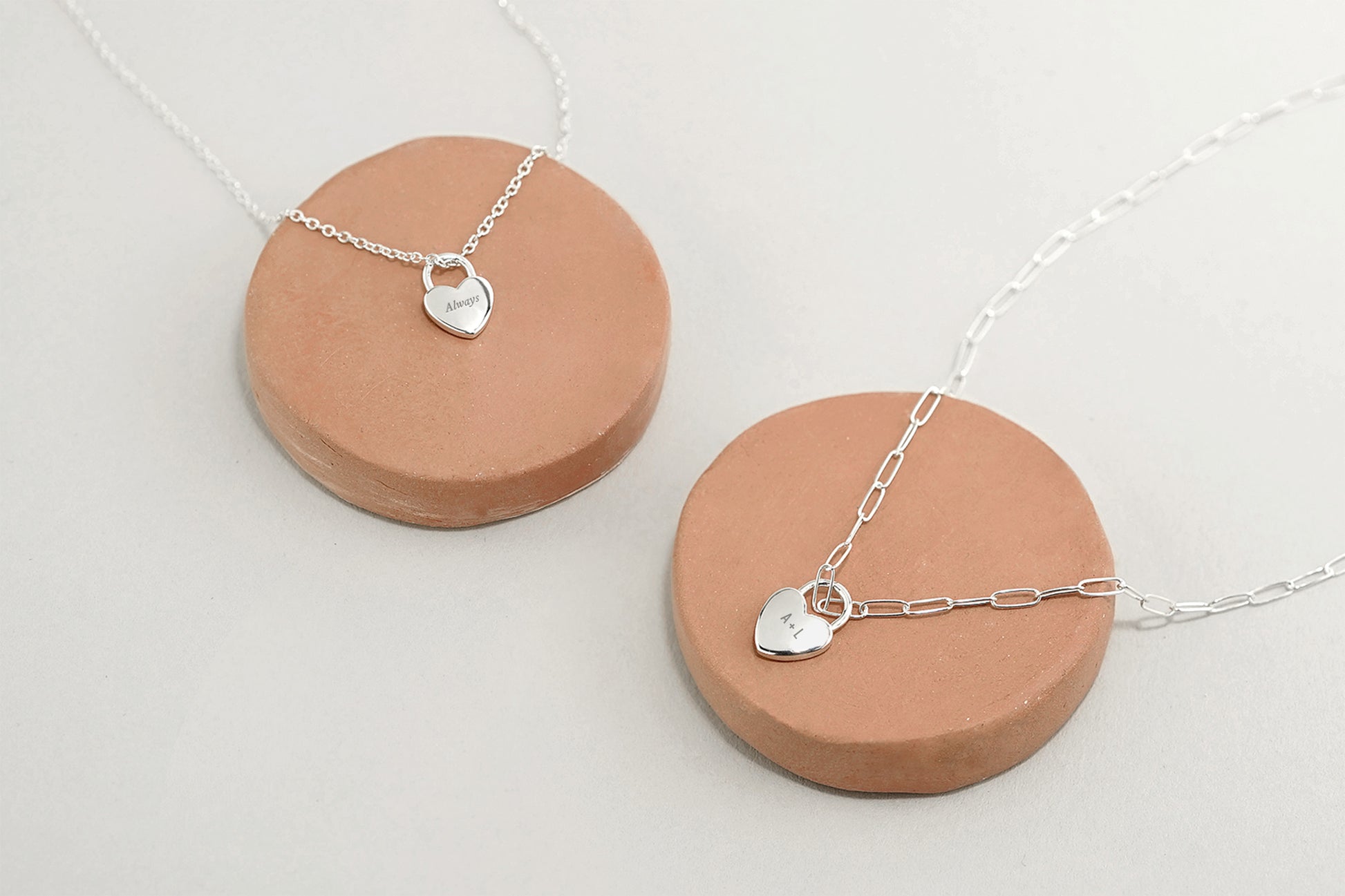 Two silver necklaces with heart pendants on ceramic stands against a light background